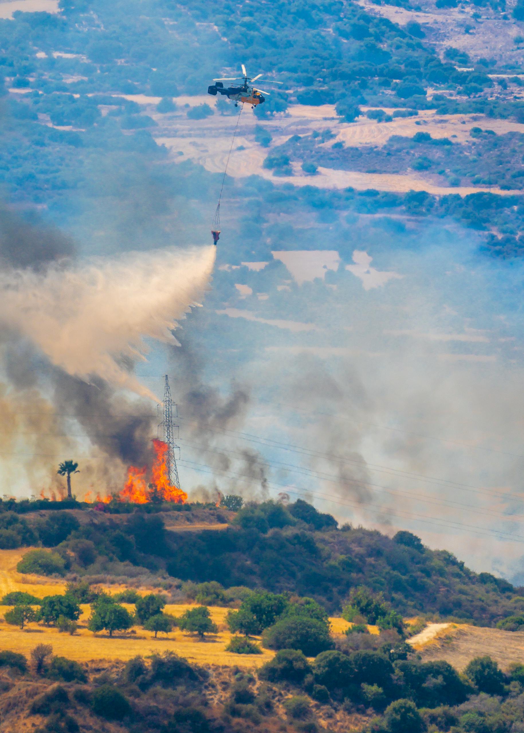 Aerial view of helicopter dousing forest fire with water bucket, showcasing fire control efforts.